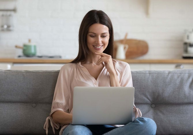 Mulher sentada na frente do computador de camisa rosa no sofá cinza analisando detalhes da sua Loja Dropshipping com ajuda do Mercado Pago