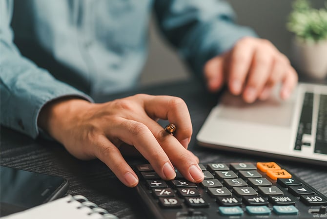 Mãos de um homem vestindo camisa social azul mexendo em um laptop e fazendo contas na calculadora, posicionada em uma mesa de escritório com caderno e celular, além de pequeno vaso de flores, e estando em busca de informações precisas sobre Linha de Crédito para MEI do Mercado Pago.