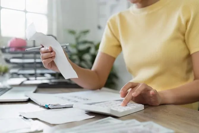 Mercado Pago: mãos de uma mulher vestindo camiseta amarela apoiadas em uma mesa com papéis e calculadora, enquanto ela segura uma conta para verificar o fluxo de caixa do seu negócio.