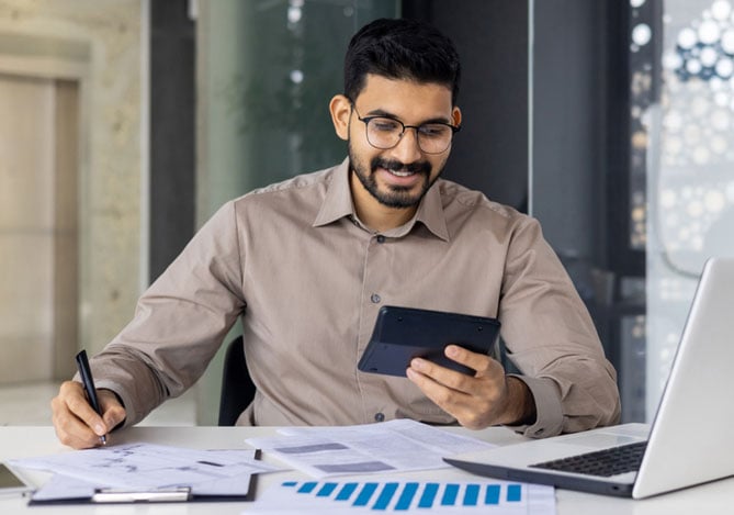 Mercado Pago: imagem de um empreendedor sentado à mesa de trabalho olhando seu celular e fazendo anotações. Sobre a mesa há alguns relatórios impressos e um notebook aberto, no qual ele verifica qual a viabilidade de fazer antecipação de recebíveis em sua loja. 