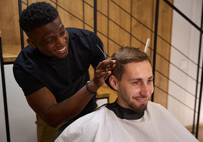 Mercado Pago: Homem sorrindo com camisa preta em pé, dono de uma barbearia por assinatura, realizando corte de cabelo em outro homem sentado em sua frente com avental branco.