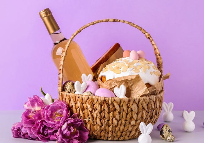 Cesta de Páscoa com doces, ovos de chocolate, bolo decorado e uma garrafa de vinho, disposta sobre uma mesa com fundo lilás, representando um presente temático para a data.