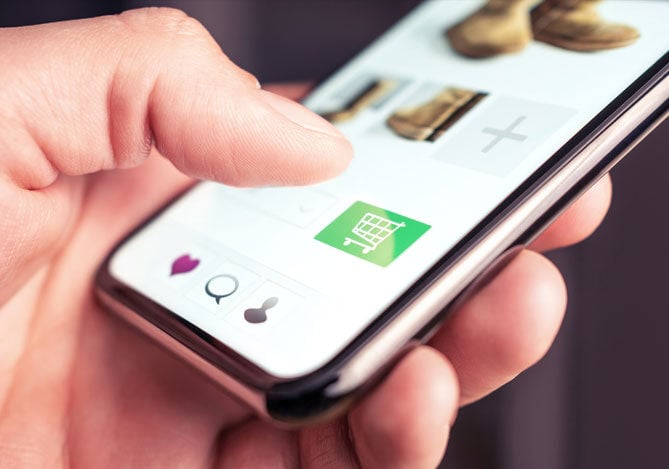 Mercado Pago: imagem da mão de uma pessoa segurando um celular enquanto realiza a compra de produtos que estão em seu carrinho em um site. Na imagem é possível ver a foto de botas e o ícone de um carrinho de compras, indicando que o site possui estratégias que otimizam o checkout para dispositivos móveis a fim de agilizar o processo de compra de seus calçados.