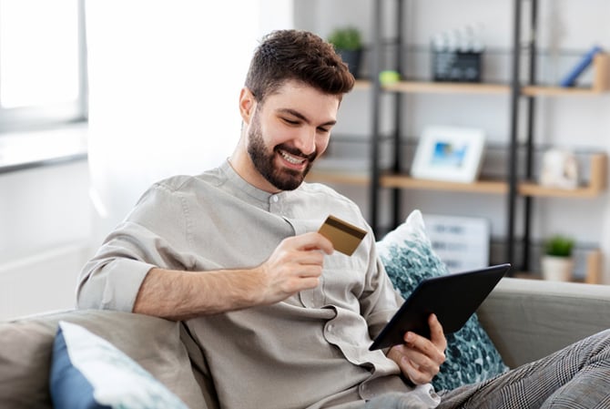 Mercado Pago: homem sorrindo sentado no sofá da sala, segurando um tablet e um cartão para realizar sua compra recorrente.