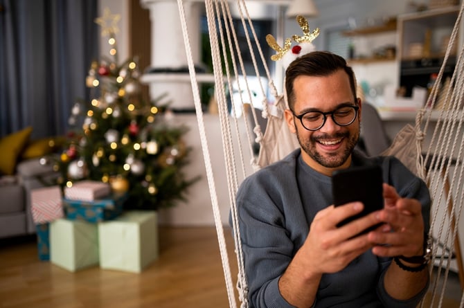 Homem de óculos, vestindo um moletom e sentando em uma rede na sua sala, com a árvore de natal atrás dele, e mexendo no celular enquanto faz compras de Natal com ajuda do Link de Pagamento do Mercado Pago