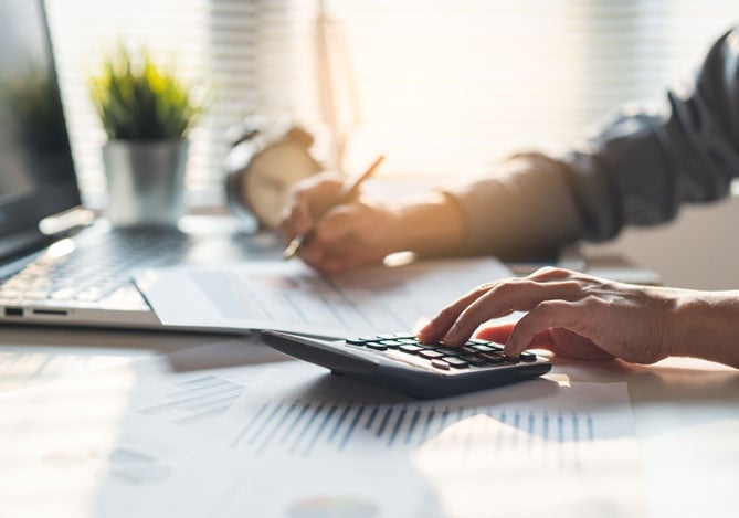 Mercado Pago: imagem das mãos de uma pessoa que empreende apoiadas sobre a mesa de trabalho, enquanto a pessoa realiza cálculos para o pagamento de débitos atrasados do DAS-MEI. Na imagem, é possível ver um notebook aberto sobre a mesa. Também é possível ver que a pessoa está fazendo anotações em um papel com uma das mãos enquanto digita em uma calculadora com a outra mão.