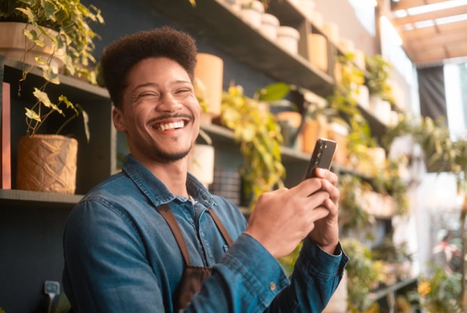 Mercado Pago: Homem sorridente usando o celular em uma loja de plantas, representando a praticidade e rapidez do Dinheiro Express Mercado Pago para gerenciar pagamentos e finanças diárias.