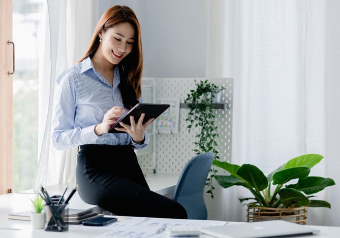 Mercado Pago: imagem de uma profissional autônoma apoiada em sua mesa de trabalho enquanto mexe em um tablet para avaliar estratégias eficientes de como conquistar a estabilidade financeira usando pagamentos recorrentes em seu negócio. Ela veste uma camisa social de cor azul clara e, sobre a mesa, é possível ver alguns papéis, um notebook fechado, canetas e, ao fundo da imagem, no chão, há um vaso decorativo com uma planta.