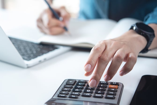 Mercado Pago: mãos de uma pessoa apoiada em mesa, a esquerda teclando em calculadora e a direita anotando em caderno. Também tem um notebook em cima dessa mesa, representando o planejamento de um empreendedor para o período de alta demanda. 