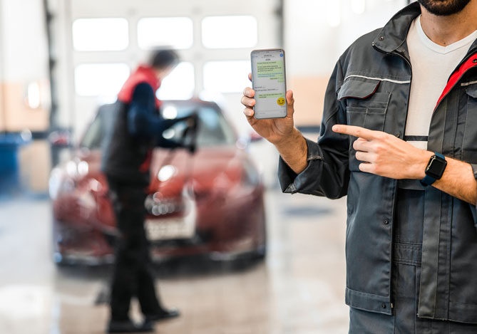 Mercado Pago: homem vestindo jaqueta preta e camiseta branca, segurando celular mostrando atendimento do lava rápido em WhatsApp. Atrás, há um outro homem de jaqueta e calça preta, lavando um carro vermelho. 