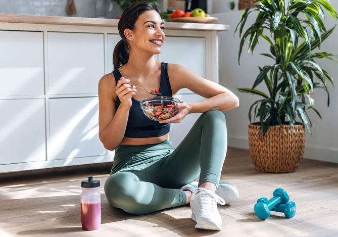 Mercado Pago: imagem de uma mulher sentada no chão de sua casa se alimentando com uma salada de frutas. Ao lado dela, no chão, é possível ver uma garrafinha e halteres adquiridos em uma loja de suplementos e artigos esportivos.