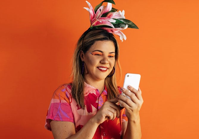 Mulher sorrindo vestindo adereços de Carnaval enquanto mexe no smartphone para explorar as soluções do Mercado Pago para ter mais vendas
