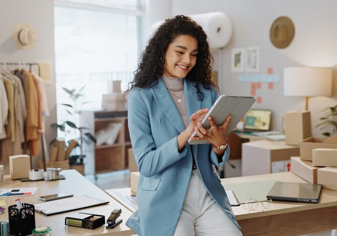 Empreendedora vestindo um paletó azul e uma calça branca, apoiada em mesa de sua loja e com um tablet em mãos, verificando como pedir empréstimo para abrir o seu negócio. Ao fundo, há algumas roupas e itens de papelaria apoiados pela mesa, além de caixas de papelão.