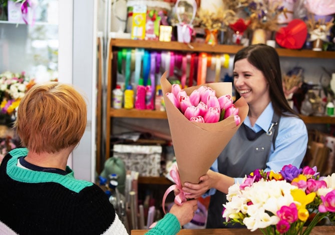 Mercado Pago: Pequenos negócios no Dia das Mães: florista sorridente entrega buquê de tulipas rosa a cliente em loja decorada com flores e fitas coloridas.