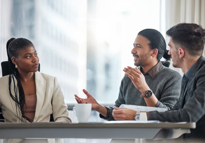 Mercado Pago: Mulher sentada em mesa de reunião, vestindo um paletó branco e uma camiseta bege, conversando sobre pitch de vendas com mais dois homens que também se sentam à mesa, vestindo blusas cinzas. Na mesa, há papéis e canecas. 