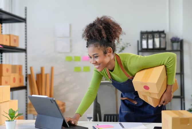Mercado Pago: mulher se apoiando em bancada para mexer no computador, dentro do seu galpão de estoque, com duas caixas sob o braço, verificando o envio de itens para clientes do plano recorrente. 