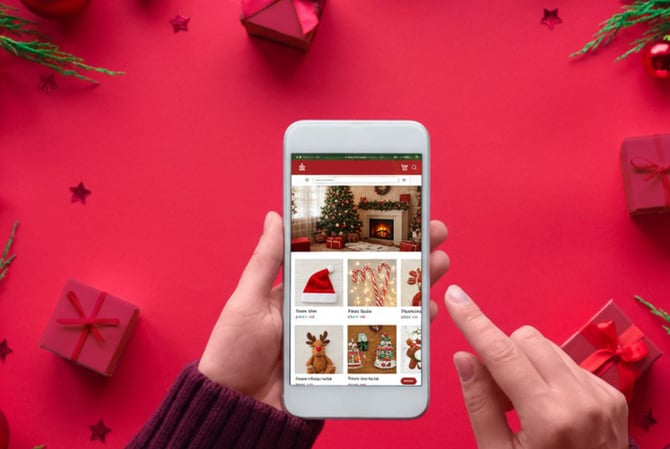 Mercado Pago: mãos segurando um celular visto de cima em frente a um fundo rosa com presentes. A tela está em um e-commerce de produtos de Natal para comprar online. 