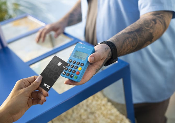 Imagem de um empreendedor recebendo um pagamento por aproximação com uma Point Mini NFC 2, ilustrando que ele teria adquirido a maquininha com um revendedor do Programa de Revenda Renda na Mão Mercado Pago, que entrou no programa como uma forma de manter uma renda extra. Na imagem, é possível ver a mão da pessoa que realiza o pagamento para o empreendedor e a mão do empreendedor segurando a maquininha. O pagamento é realizado com um Cartão Mercado Pago.
