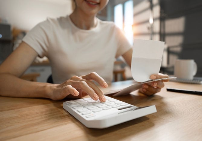 Mercado Pago: imagem de uma pessoa usando uma calculadora branca para avaliar as finanças do seu negócio a fim de criar uma reserva financeira como segurança. Na imagem, é possível ver que a calculadora está apoiada sobre uma mesa e a pessoa verifica  notas e contas para fazer uma análise completa.
