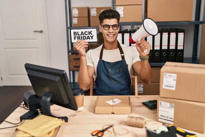 Mercado Pago: empreendedor sentado à mesa, com caixas, uma tela, um impressora, tesoura, fita e estilete espalhados. Atrás dele, uma prateleira com caixa e pastas. Ele está sorrindo, com uma mão segurando um megafone e a outra uma placa que anuncia a chegada da semana Black Friday.
