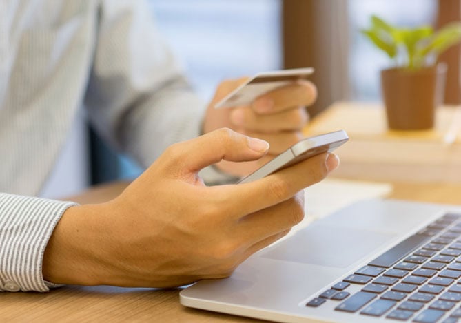 Mercado Pago: imagem das mãos de uma pessoa segurando um celular em uma das mãos e um cartão de crédito na outra para efetuar pagamentos de encomendas de Páscoa por meio de links de pagamentos enviados por empreendedores confeiteiros. Na imagem, também é possível ver que a pessoa está com as mãos apoiadas sobre uma mesa onde há um notebook aberto.