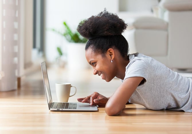 Mulher deitada com uma caneca no chão e sorrindo enquanto usa o laptop