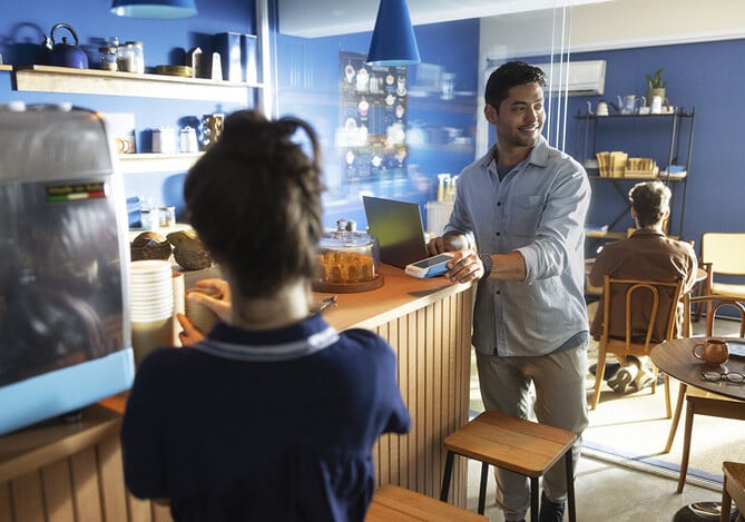 Café com paredes azuis, utensílios, mesas e clientes, e um empreendedor em pé, em frente ao laptop, vestindo calça jeans e camisa, segurando uma maquininha que receba na hora, a Point Mercado Pago, para cobrar o cliente. E uma mulher de costas preparando um pedido. 