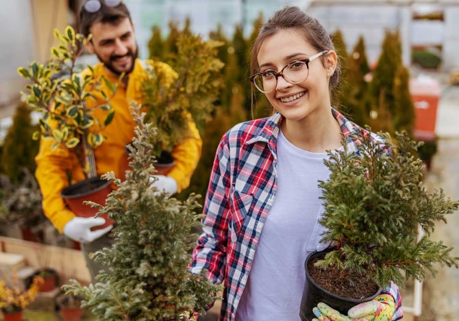 Mulher sorridente segurando dois vasos de planta e um homem no fundo também segurando dois vasos de plantas