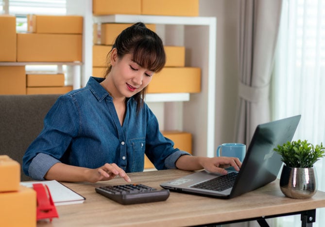 Mercado Pago: empreendedora no estoque de sua loja em frente ao computador calculando planos de assinatura