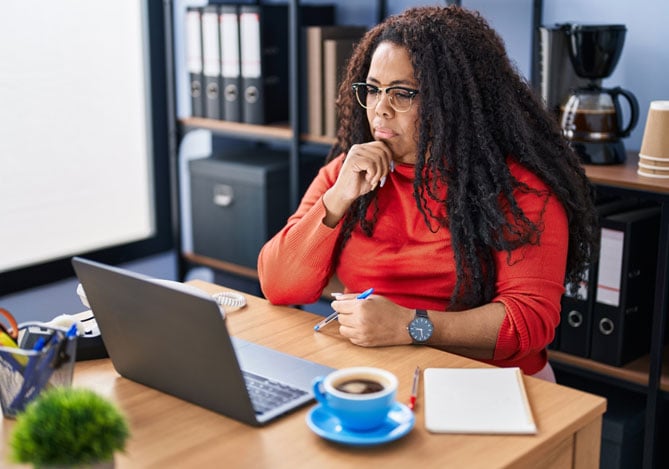 Empreendedores de blusa vermelha e óculos, com a mão no queixo sentada em sua mesa de trabalho com notebook aberto pesquisando sobre as recusas de pagamento da Point Mercado Pago