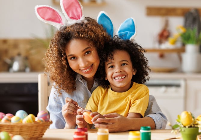 Nicho de mercado Mercado Pago: Mulher com blusa azul e criança com blusa amarela, ambos vestindo orelhinhas de coelho na cabeça, sentados em mesa de cozinha pintando ovos da cor amarela, vermelho e verde. 