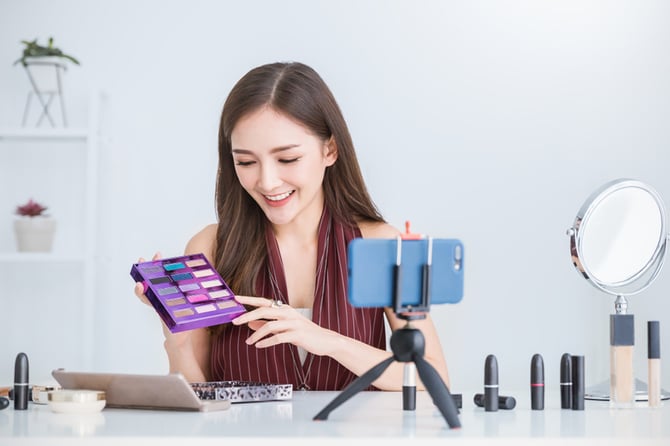 Mercado Pago: empreendedora em sala realizando Live commerce com celular apoiado em mesa e produtos de maquiagem em mãos