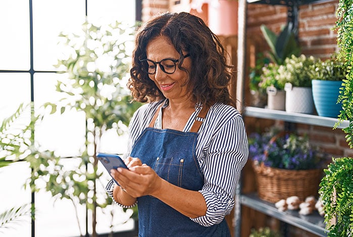 Mulher, dona de uma floricultura, em um dos ambientes do seu negócio, vestindo camisa social listrada em azul e branco, com um avental jeans por cima, mexendo no seu celular para pagar DAS MEI pelo Mercado Pago.
