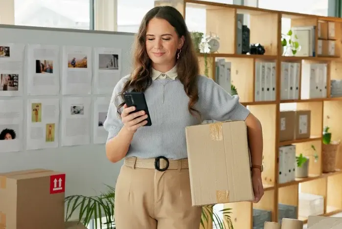 Mercado Pago: mulher em seu atelier, segurando uma caixa com um braço e um celular na outra mão, para programar a cobrança automática dos seus clientes. 