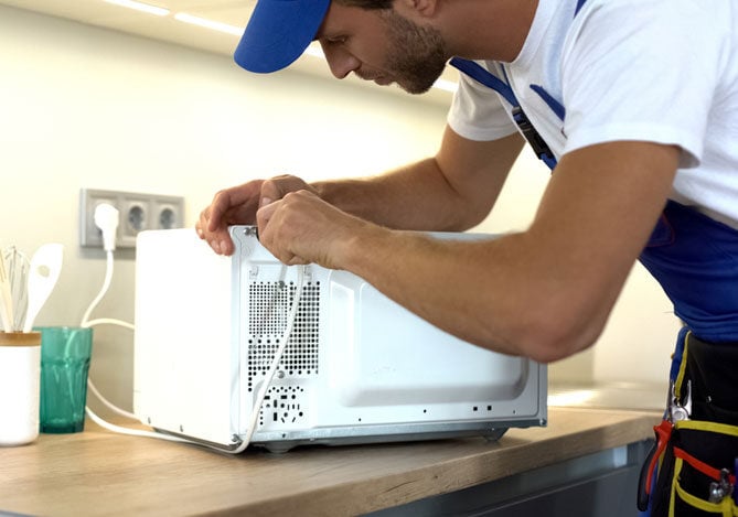 Mercado Pago: imagem de um profissional de assistência técnica vestindo macacão e utilizando equipamentos de segurança, enquanto conecta um fio na parte de trás de um micro-ondas que está apoiado em uma bancada de madeira.