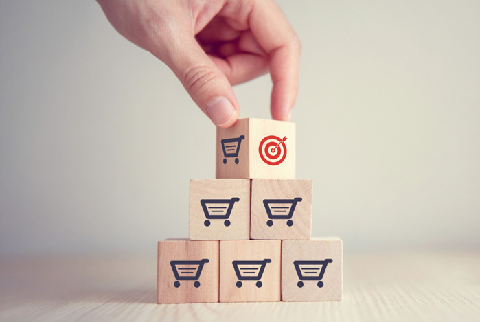 Mercado Pago: mão empilhando cubos de madeira estampados com carrinho e um alvo, formando uma pirâmide para representar uma loja tentando aumentar as vendas com promoções estratégicas.
