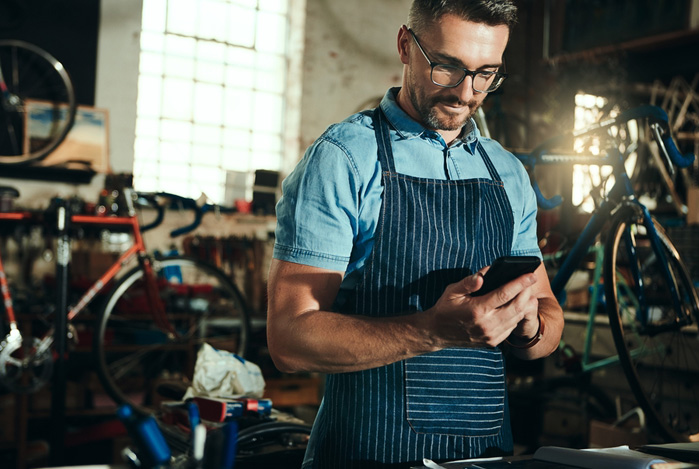 Mercado Pago: homem de óculos, usando camisa polo azul com um avental azul-escuro com listras finas em branco, em sua oficina de bicicletas mexendo no celular para avaliar como está a capital de giro do seu negócio.