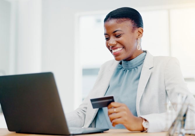Mercado Pago: mulher em escritório, sentada à mesa em frente ao notebook, sorrindo e com o cartão de crédito empresarial na sua mão. 