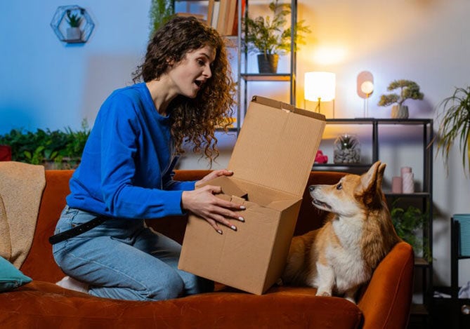 Mercado Pago: imagem de uma mulher sentada no sofá da sala de sua casa em frente ao seu cachorro de estimação, segurando uma caixa aberta que acabou de receber em um clube de assinaturas para produtos pet, do qual faz parte. 