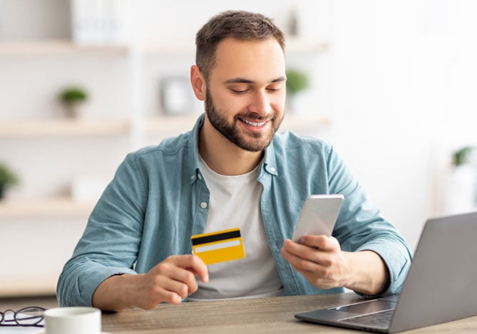 Homem vestindo camiseta branca e camisa azul por cima, sorrindo e sentado com os braços apoiados na mesa com laptop, caderno, xícara e óculos, enquanto segura o celular em uma mão e o cartão de crédito na outra para ativar uma cobrança recorrente com ajuda do Mercado Pago.