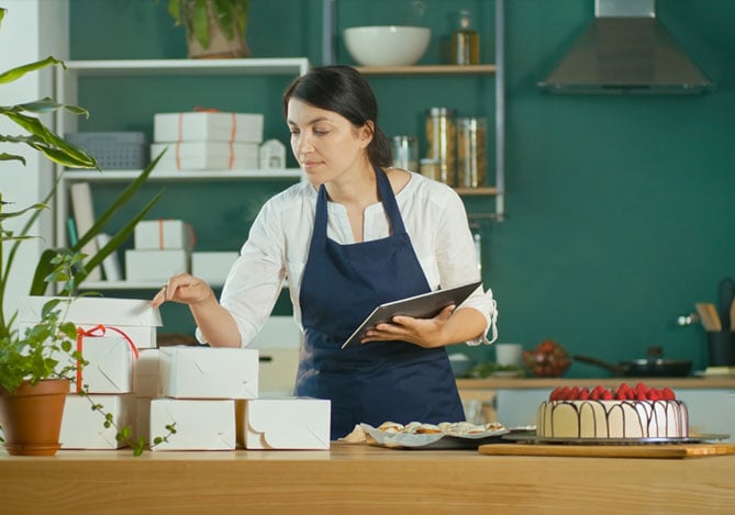 Mercado Pago: imagem de uma empreendedora de confeitaria vestindo camisa branca e avental azul em frente à sua bancada de entregas. Na imagem, é possível ver que a empreendedora está conferindo pedidos enquanto olha algumas caixas empilhadas na bancada. Em uma das mãos, a empreendedora segura um tablet para verificar as informações dos clientes que ela já organizou após verificar as melhores estratégias de como vender bolos pela internet. Sobre a bancada há um bolo confeitado com glacê branco, morangos no topo e fios de chocolate, além de um prato com docinhos, prontos para serem embalados para entrega.