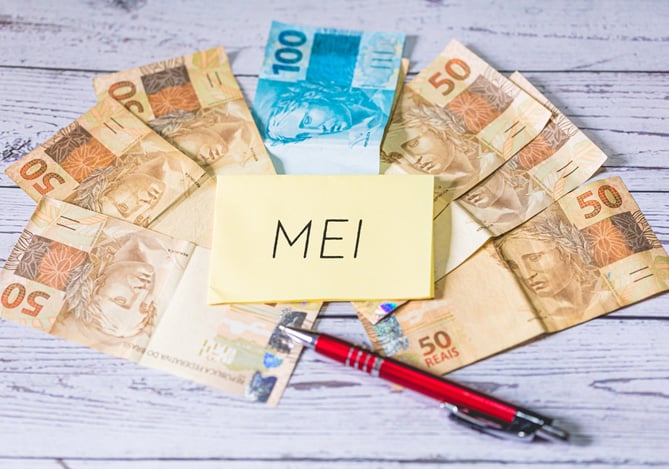 Mercado Pago: um leque com notas de cinquenta e cem reais, com uma caneta e um papel escrito “MEI” no centro, representando uma Conta MEI em instituições financeiras.