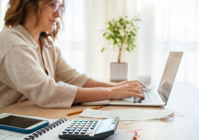 Imagem de uma empreendedora sentada à mesa de trabalho digitando em seu notebook enquanto faz o controle financeiro de seu negócio por meio do Sistema de Gestão Mercado Pago em sua Conta PJ. Sobre a mesa, é possível ver algumas folhas, um celular que ela usa para fazer o acompanhamento em tempo real das finanças do seu negócio via app Mercado Pago e uma calculadora.