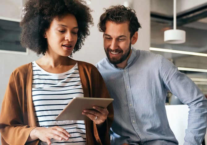 Mercado Pago: imagem de dois empreendedores no escritório do negócio discutindo estratégias de vendas. Na imagem, é possível ver uma mulher segurando um tablet em uma das mãos, enquanto o homem, ao seu lado, olha para a tela e sorri. Ao fundo da imagem, há bancadas de trabalho.