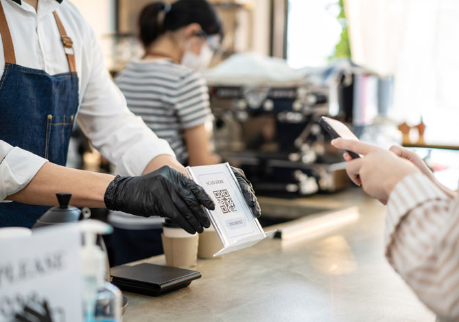 Mercado Pago: Vendedor de um café segurando uma placa com Código QR enquanto a cliente faz a leitura com o smartphone para fazer seu pagamento