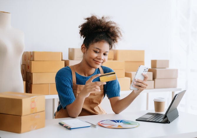 Mercado Pago: Empreendedora vestindo camiseta azul e avental amarelo, segurando cartão e celular pensando em formas de realizar a fidelização de clientes com vendas recorrentes. Ela está apoiada em mesa com notebook e caderno, e há algumas caixas atrás. 