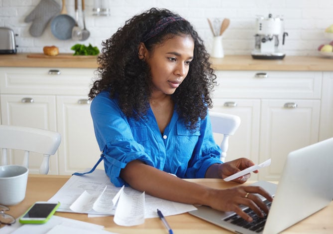 Mulher com camisa azul sentada na mesa da cozinha e mexendo em um laptop enquanto segura o smartphone com a outra mão para analisar suas finanças pessoais e corporativas com ajuda do Mercado Pago.