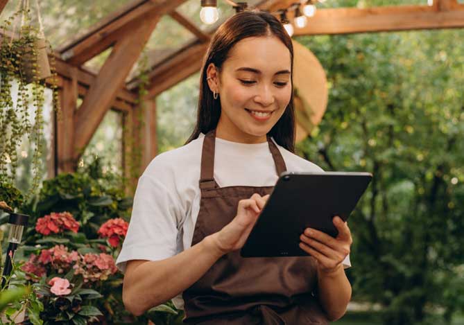 Mercado Pago: imagem de uma empreendedora em um espaço rodeada por plantas e flores, vestindo camiseta branca e avental, enquanto confere informações sobre seu fluxo de caixa em um tablet.