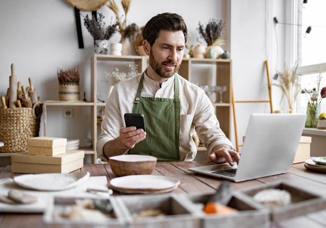 Mercado Pago: Homem com avental verde trabalhando em casa, usando celular e notebook em uma mesa de artesanato, representando uma nova fonte de renda com criatividade e autonomia.