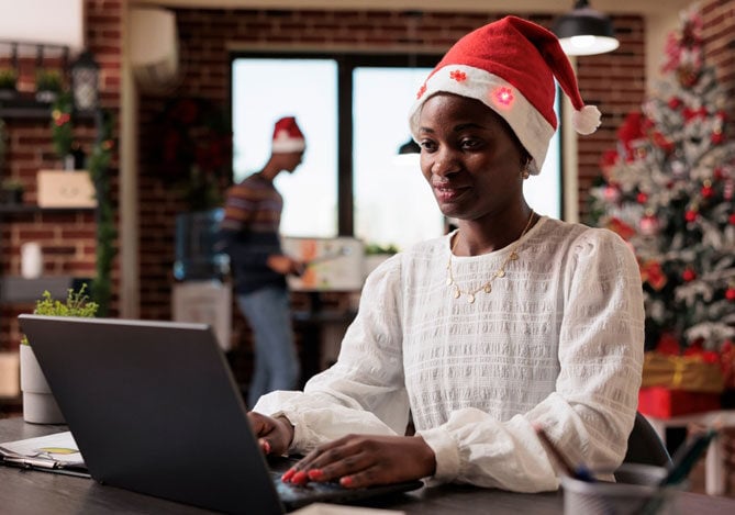 Mercado Pago: imagem de uma empreendedora vestindo uma blusa de manga longa branca e gorro de Natal sentada à mesa de trabalho enquanto digita em seu notebook para fazer a gestão de vendas de sua loja. Ao fundo, é possível ver uma pessoa que também está no escritório da loja, que está enfeitada para as festas de fim de ano.